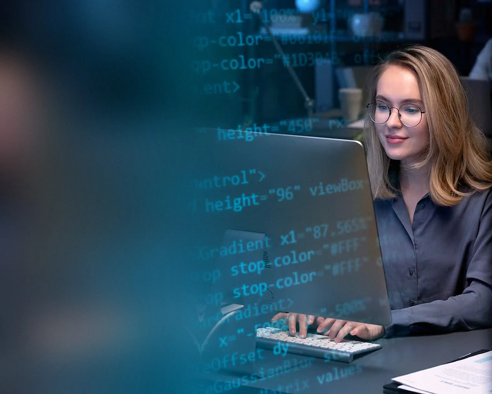 Software developer smiling at her computer while lines of code glow across the glass in front of her